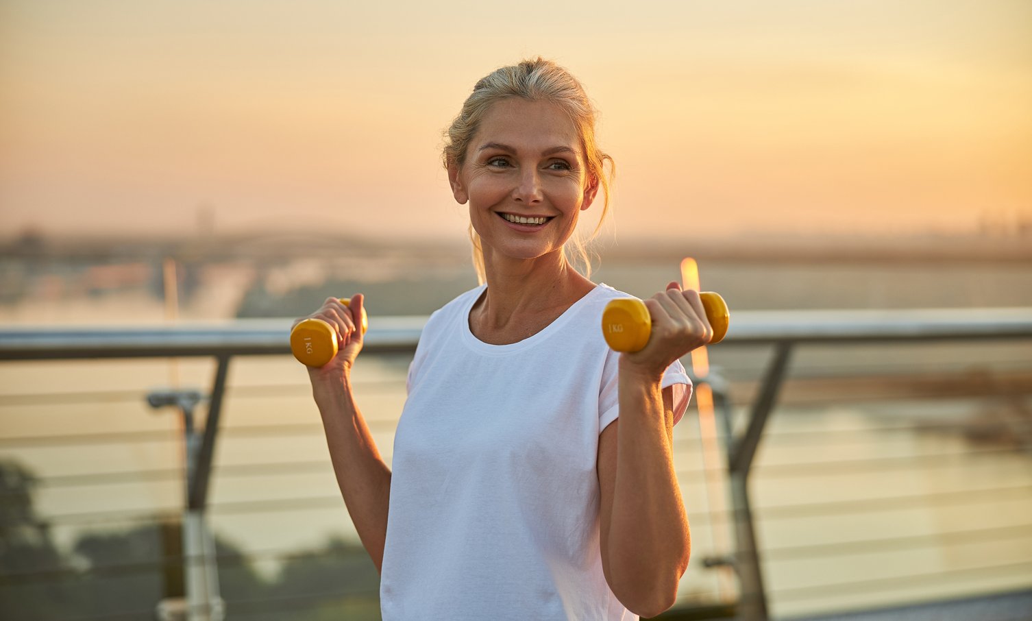Smiling middle-aged woman exercising with yellow dumbbells outdoors at sunrise on a balcony overlooking water.