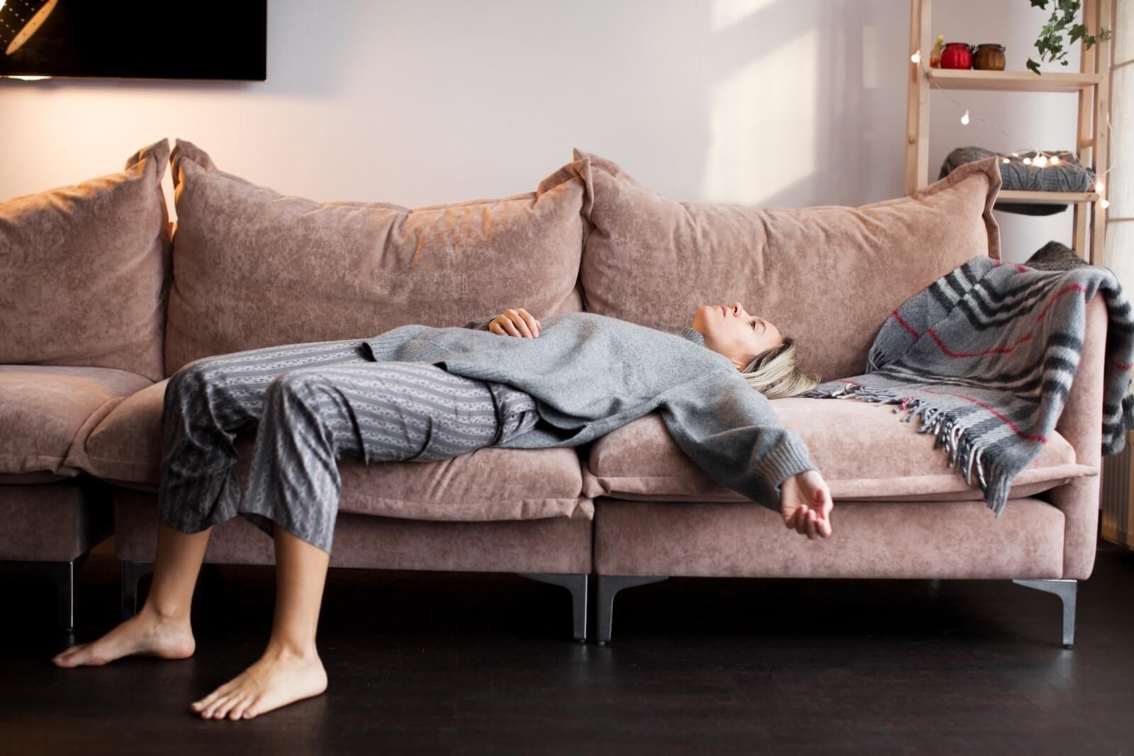 Tired woman in loungewear lying sprawled on a beige sofa in a cozy living room, appearing exhausted or overwhelmed.