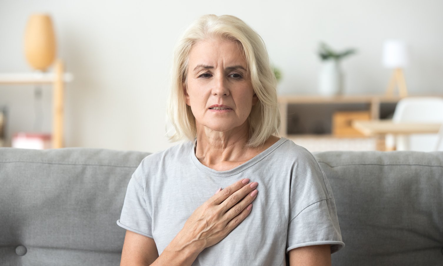 Concerned older woman sitting on a couch with her hand on her chest, appearing to experience discomfort or pain in a calm home setting.