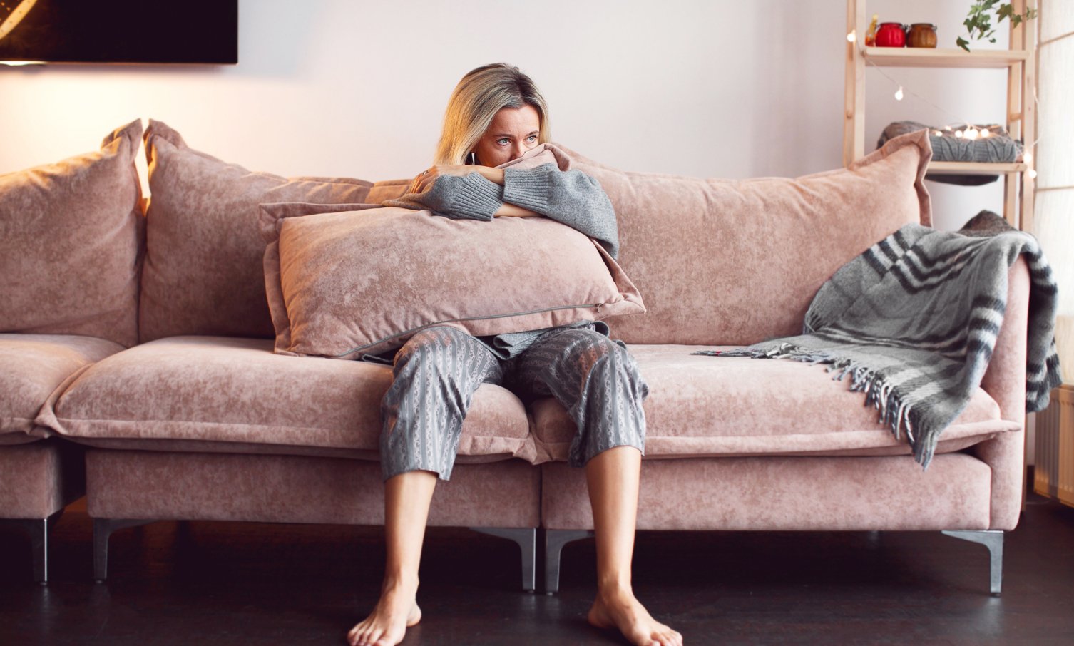 Anxious-looking woman in loungewear sitting on a sofa, hugging a large pillow with a worried expression.