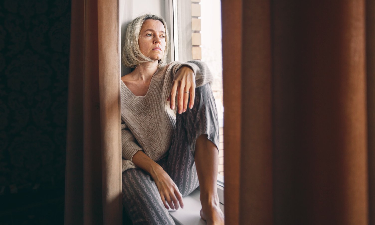 Thoughtful middle-aged woman sitting by a window in loungewear, looking out with a pensive expression, partially framed by curtains.