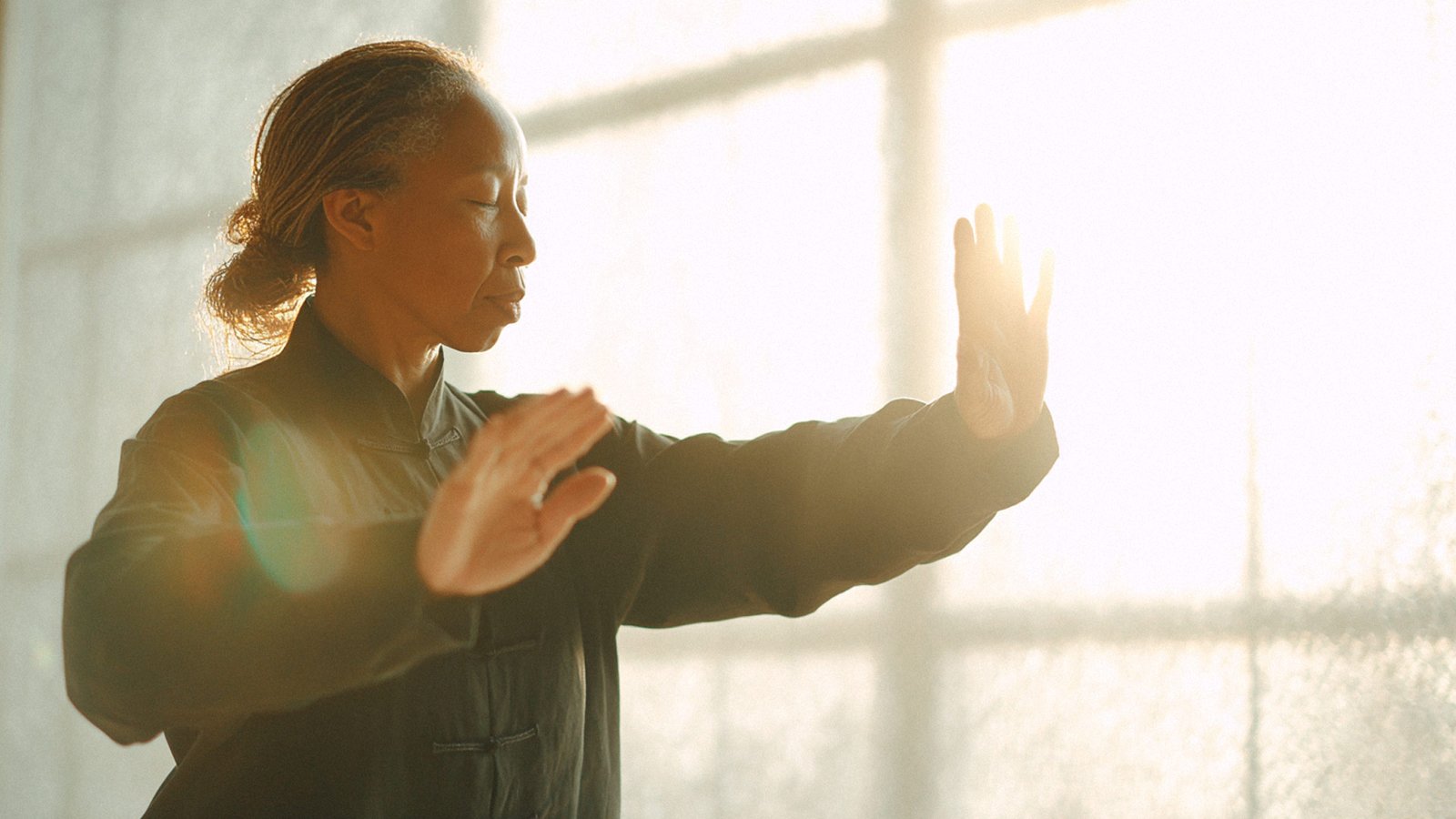 A woman practicing tai chi with eyes closed, hands raised in a calm, focused movement, bathed in warm sunlight.