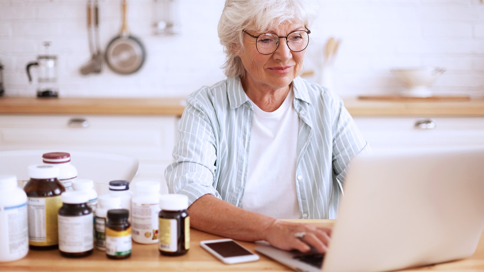 An older woman with white hair and glasses sits at a kitchen table using a laptop, surrounded by various supplement bottles, appearing to research or compare products.