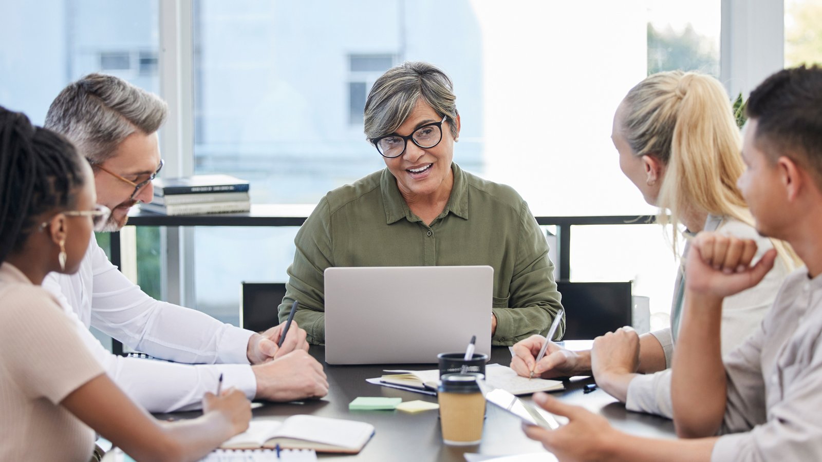 A middle-aged woman with short gray hair and glasses leads a meeting at a table, speaking while working on a laptop, surrounded by colleagues taking notes.