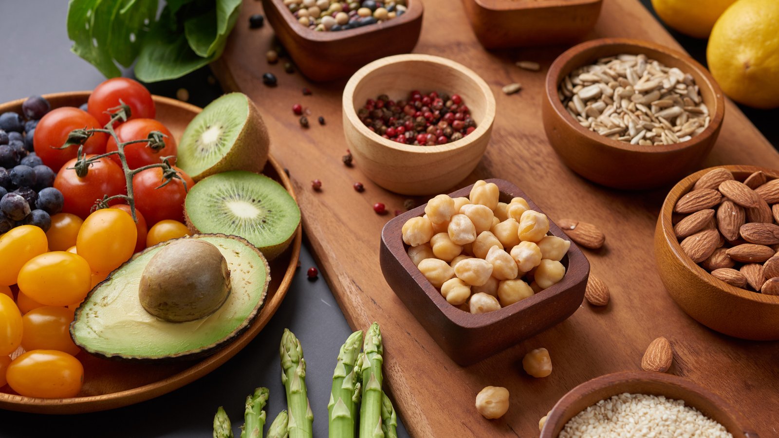 Close-up of colorful high-fiber foods, including tomatoes, blueberries, kiwi, avocado, asparagus, chickpeas, nuts, seeds, and spices arranged on a wooden board.