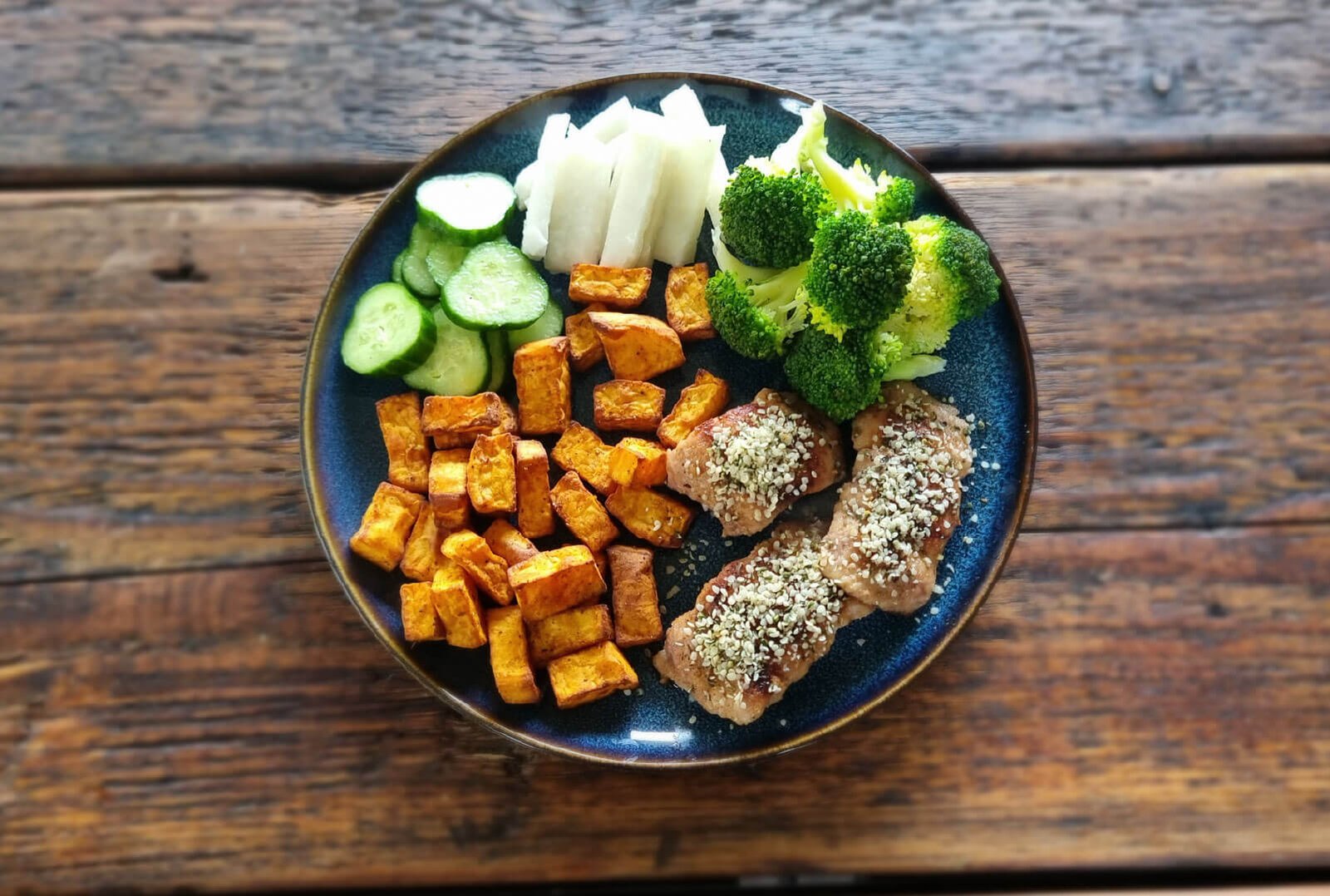 A bowl filled with sliced cucumbers, raw white vegetable sticks, steamed broccoli, roasted sweet potato cubes, and seared catfish topped with seeds.
