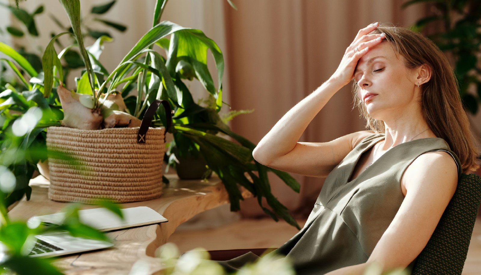 Woman sitting at a desk in a sunlit room, eyes closed and hand pressed to her forehead, appearing fatigued or overwhelmed; surrounded by houseplants with an open laptop nearby.